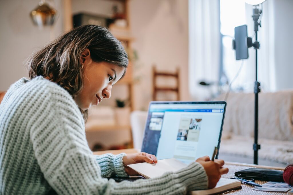 Girl working from home on laptop, taking notes and recording via smartphone